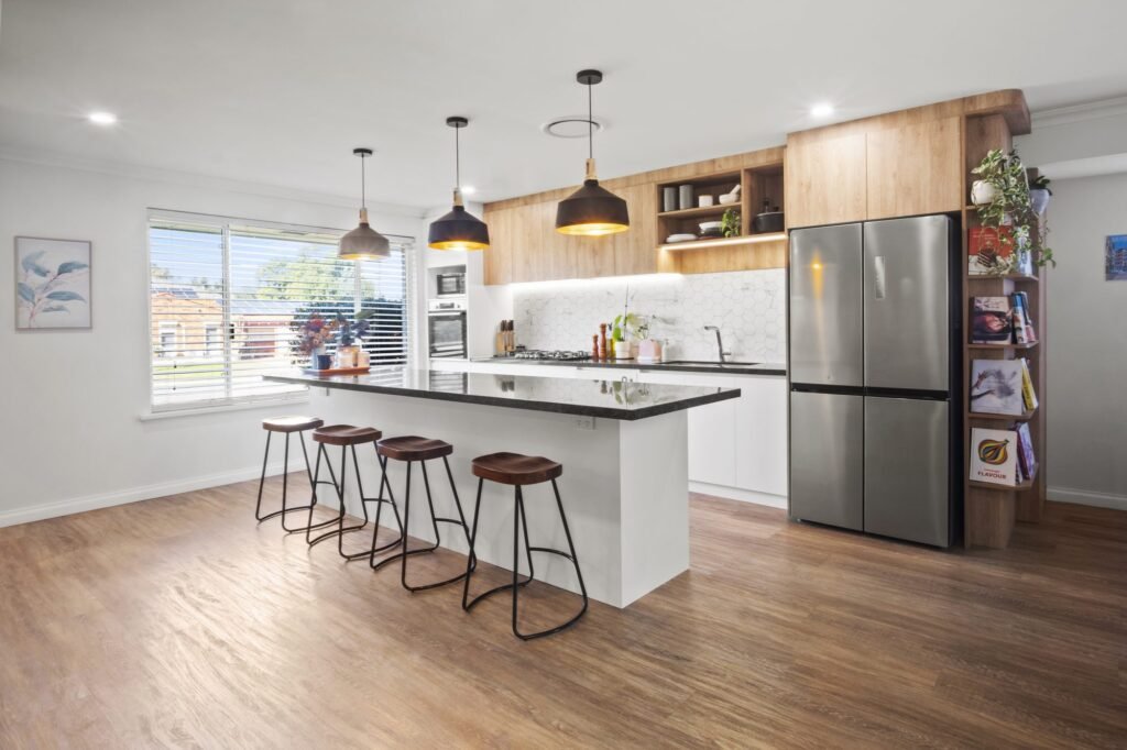 The island from the living side: four timber-seat stools, three black pendants, oak-look overheads and the stainless fridge with cookbook shelves on its end