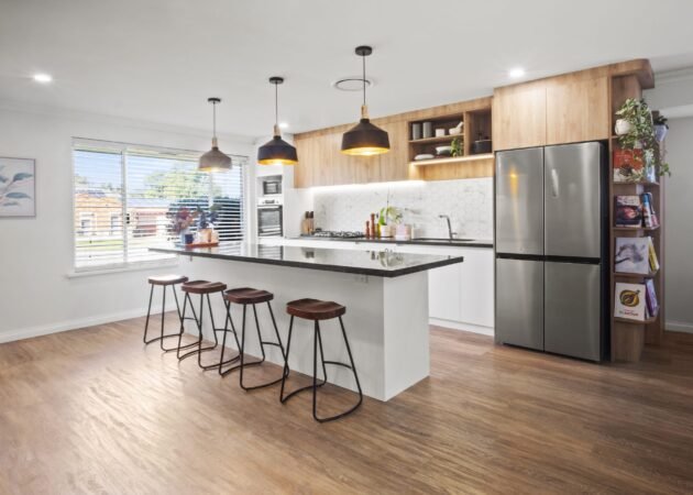 The island from the living side: four timber-seat stools, three black pendants, oak-look overheads and the stainless fridge with cookbook shelves on its end