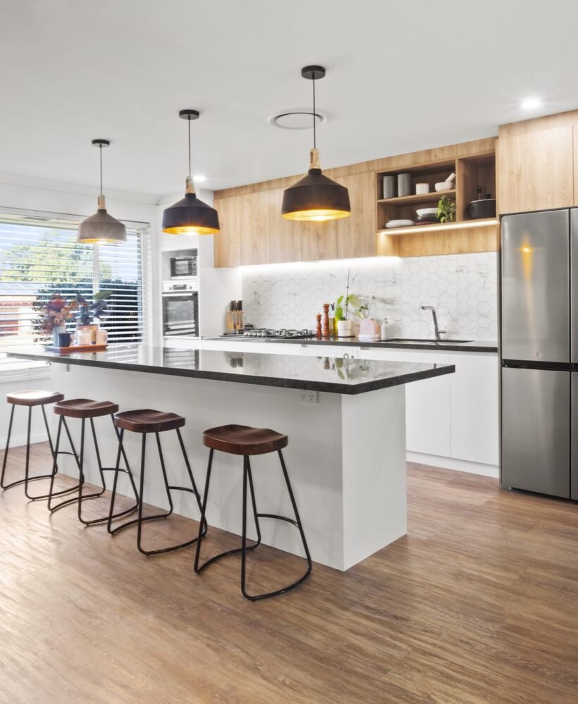 The island from the living side: four timber-seat stools, three black pendants, oak-look overheads and the stainless fridge with cookbook shelves on its end