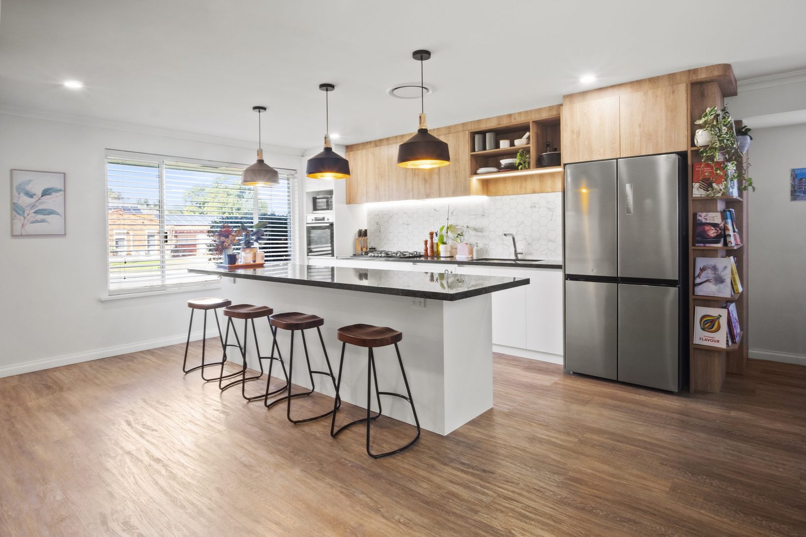 The island from the living side: four timber-seat stools, three black pendants, oak-look overheads and the stainless fridge with cookbook shelves on its end